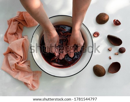 Woman's hand soaking fabric in pot with natural avocado dye, ingredients and pink textile as end result. Royalty-Free Stock Photo #1781544797