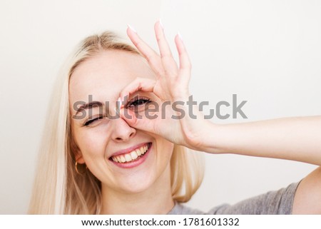 Close up portrait of beautiful joyful blonde female smiling, demonstrating white teeth, looking at the camera through fingers in okay gesture. Face expressions, emotions, and body language