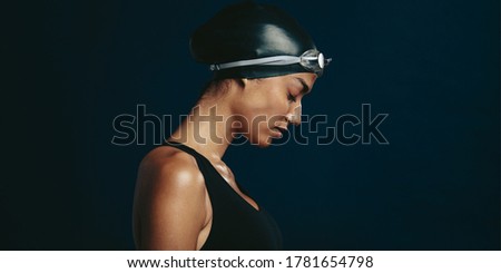 Side view of a professional female swimmer with goggles and a hat. Woman wearing black swimsuit, swimming cap and goggles on dark background looking exhausted. Royalty-Free Stock Photo #1781654798