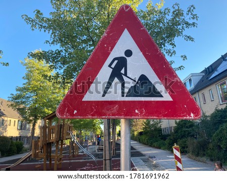 Road sign - repair work. White sign with red frame. In the background you can see a playground, green trees and houses. Summer sunny day.