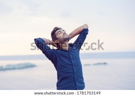 Man smiling looking up to blue sky taking deep breath celebrating freedom sea background at sunset. Positive emotion face expression feeling success peace mind concept. Free happy guy enjoying nature Royalty-Free Stock Photo #1781703149