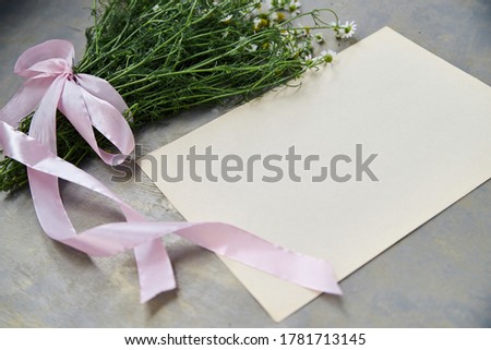 Small camomiles bouquet tied with pink ribbon and beige paper sheet on grey wooden table. Natural background picture of wild flowers. Ecological texture for poems, letters, romantic notes.