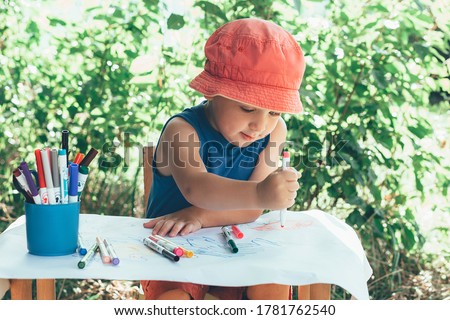 a boy in red Panama sits in nature at a children's table and draws markers on a white watman