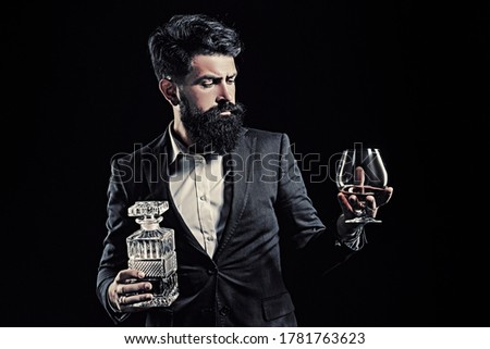 Bartender leather apron holding brandy snifter. Hipster with beard and mustache in suit drinks alcohol after working day