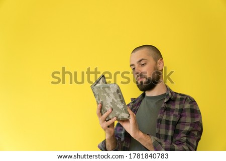 A young caucasian man with a beard on a yellow background is reading a book in a military cover. Focused an serious guy with copy space