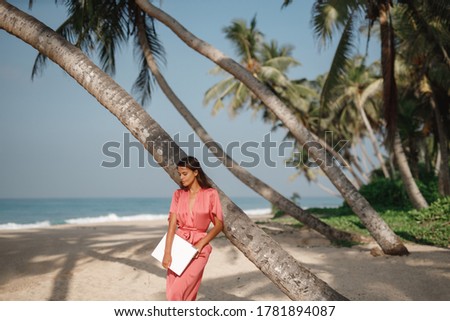 Young woman in a hat working with laptop computer on tropical island beach under palm trees Freelance concept