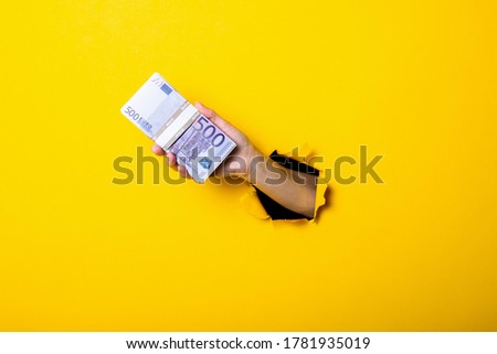 Woman's hand holds a bundle of five hundred euro bills on a yellow background.