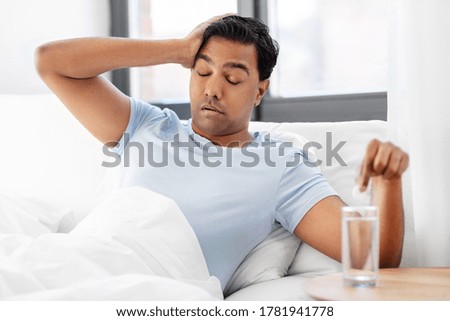 people, bedtime and rest concept - sick indian man lying in bed and dropping medicine pill into glass of water at home