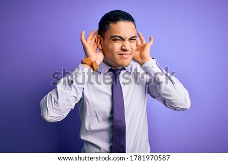 Young brazilian businessman wearing elegant tie standing over isolated purple background Trying to hear both hands on ear gesture, curious for gossip. Hearing problem, deaf