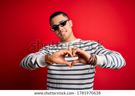 Young brazilian man wearing funny thug life sunglasses over isolated red background smiling in love showing heart symbol and shape with hands. Romantic concept.