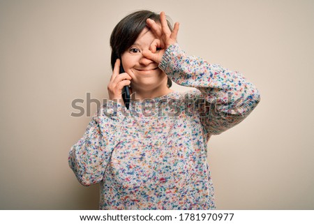 Young down syndrome woman having a conversation speaking on smartphone with happy face smiling doing ok sign with hand on eye looking through fingers