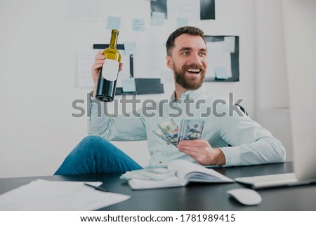 bearded man drinking wine celebrating successful business deal in modern office holding cash in his hand like card deck looking excited, money making motivation concept