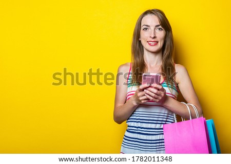 Beautiful young woman holding a phone in her hands, with packages with purchases on a yellow background.