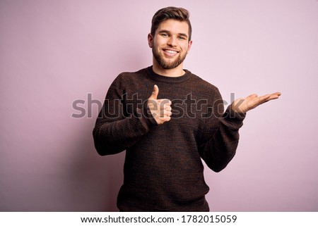 Young blond man with beard and blue eyes wearing casual sweater over pink background Showing palm hand and doing ok gesture with thumbs up, smiling happy and cheerful