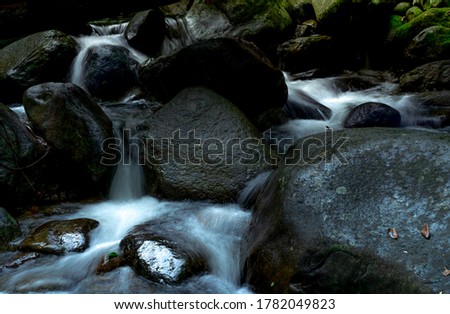 Black rock at waterfall with green moss and sunshine on rock. Waterfall is flowing in jungle. Waterfall in tropical forest. Nature background. Green season travel. Eco tourism. Waterfall in wild.