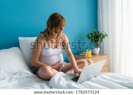 A smiling and happy pregnant caucasian woman in a sitting position in her bed while working on her laptop. One hand is on her belly.