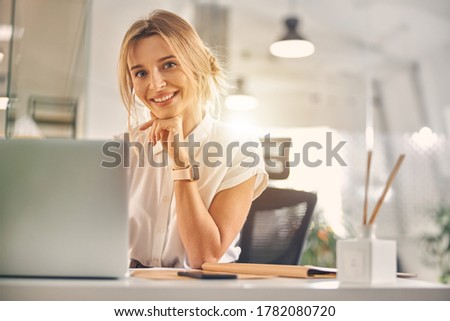 Charming blonde lady looking at camera and smiling while sitting at the table with laptop at work