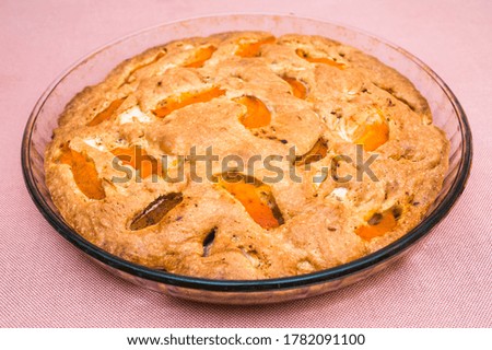 A large, round baked apple and apricot pie stands in a glass form on a table with a pink tablecloth. Delicious, fresh dessert.
