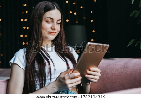 Beautiful young smiling caucasian woman resting on a pink sofa and using tablet computer. Chatting with friends, browsing information, studying online.