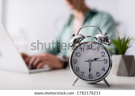 Selective focus of alarm clock on table near man using laptop at home, concept of time management Royalty-Free Stock Photo #1782178211