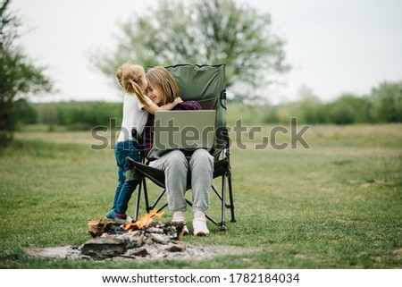 Mother work on Internet with kid outdoors. Quarantine, closed nursery school during coronavirus outbreak. Communication with family online on laptop near fire in nature. Homeschooling, freelance job.
