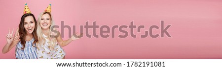Two cheerful cute young women in hats hugging and celebrating birthday over pink background
