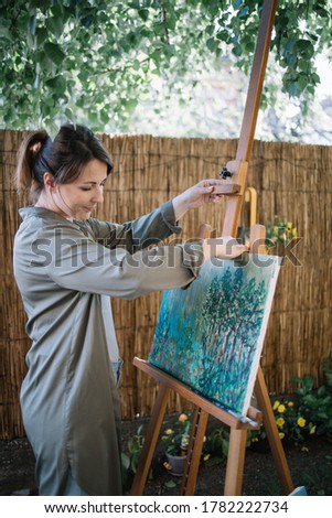 Girl getting canvas from tripod in garden. Woman's hand holding painting and canvas stand while standing in backyard with flowers and plants.