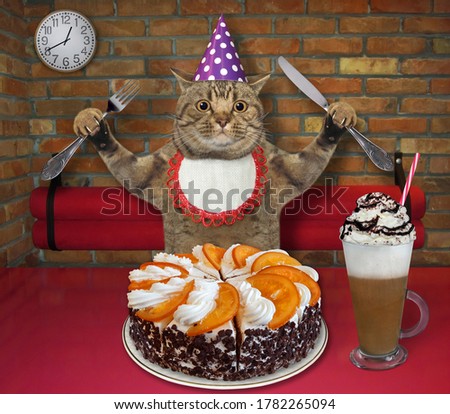 The beige big eyed cat in a party hat with a fknife and a fork in his paws is eating a holiday orange cake and drinking coffee with whipped cream at a table in a restaurant.
