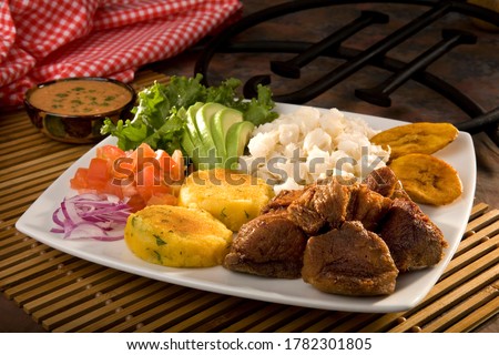 Traditional and famous Ecuadorian dish called "Fritada". It's a braised pork dish that is served always with potato fried cakes, cooked corn kernels with egg, avocado, plantains, tomato and onions. Royalty-Free Stock Photo #1782301805
