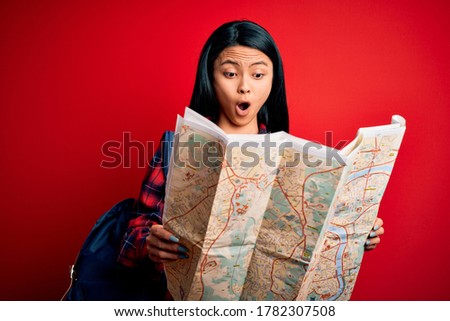 Young beautiful chinese tourist woman holding city map over isolated red background scared in shock with a surprise face, afraid and excited with fear expression