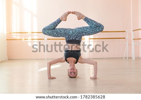 Flexible Caucasian girl stands on her head in a dance class. The girl does yoga. . Woman dancing breakdance on the background of ballet barre.