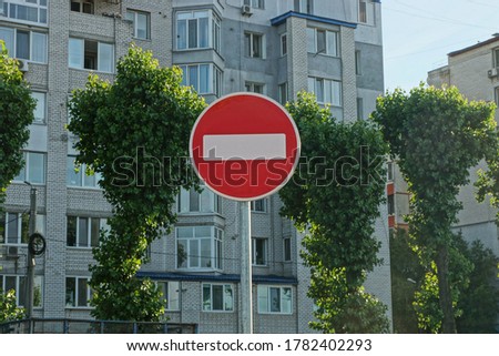 one round road sign no entry on a gray pillar against the background of green trees and a multi-storey building