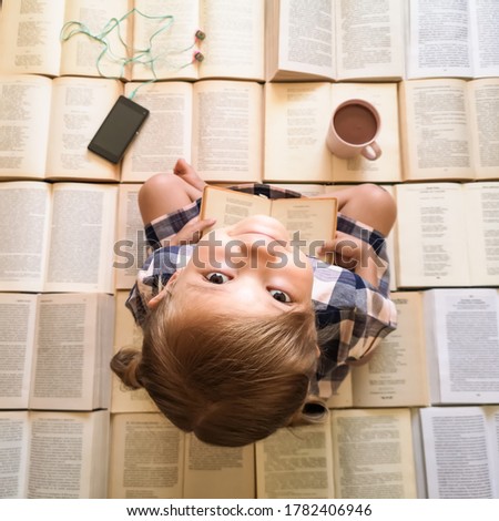 A little cute girl sits on open books, smiles and holds a book in her hands, next to it there is a pink mug with cocoa and a phone with headphones. Concept: love of reading in children, love of books