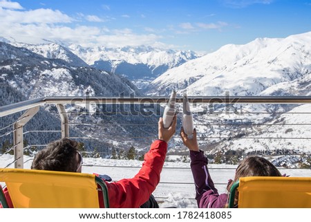 Couple in winter clothes sitting on sun loungers on a balcony toasting a bottle of beer overlooking a snowy valley
