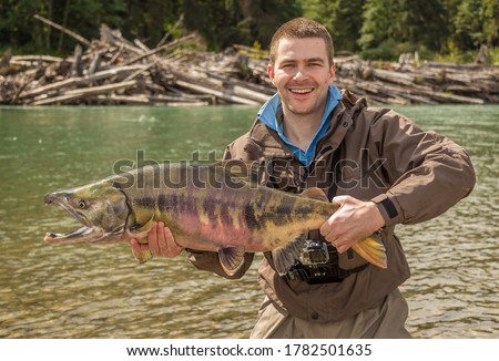 A happy sport fisherman holding up a big trophy Chum Salmon, with a log jam in the background, on the glacial green Kitimat River, British Columbia, Canada Royalty-Free Stock Photo #1782501635