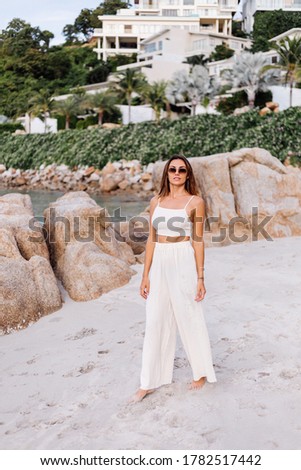 Romantic portrait of young calm happy caucasian fit slim woman in crop cami top and pants set sits alone on rocky tropical beach at sunset. Luxury villas on background. 