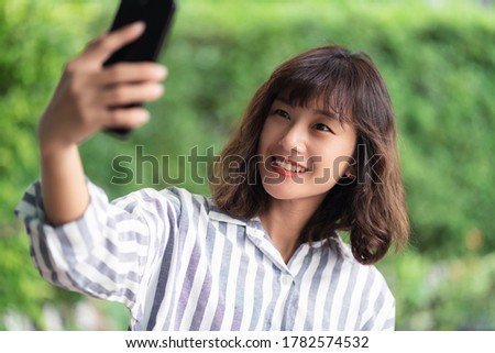 Young beautiful Asian woman using smartphone taking selfie photo. A happy girl enjoying outdoor lifestyle surrounded by greeny plant. Positive thinking concept. Bangkok, Thailand