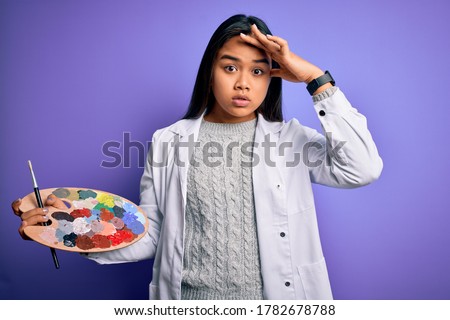 Young asian artist girl painting using paintbrush and palette with colors over purple background stressed with hand on head, shocked with shame and surprise face, angry and frustrated. Fear and upset