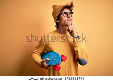 Young handsome student man holding skate wearing glasses over isolated yellow background serious face thinking about question, very confused idea