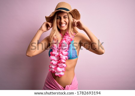 Young beautiful blonde woman on vacation wearing bikini and hat with hawaiian lei flowers covering ears with fingers with annoyed expression for the noise of loud music. Deaf concept.