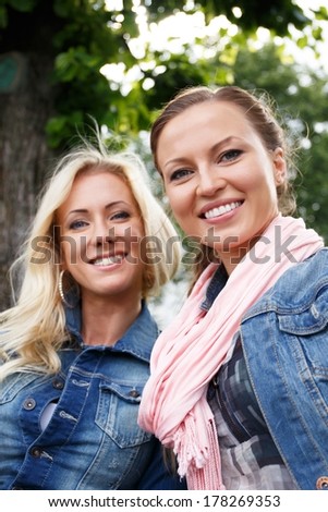 Two beautiful smiling young women in jeans jackets sitting on a bench in a park