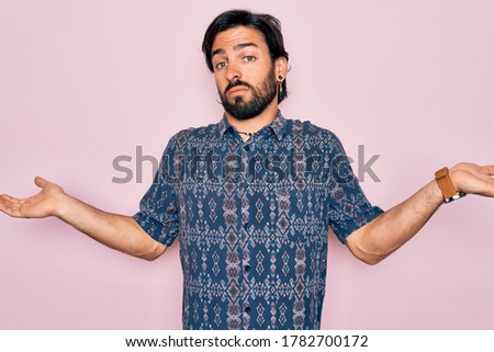Young handsome hispanic bohemian man wearing hippie style over pink background clueless and confused expression with arms and hands raised. Doubt concept.