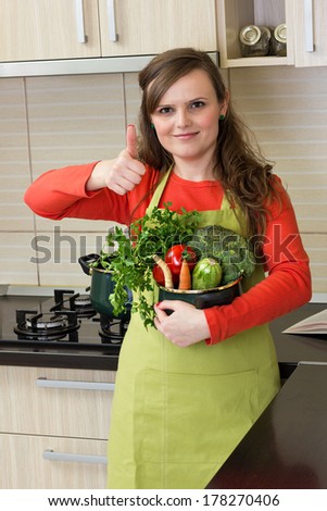 Beautiful happy young woman with apron holding a pot and making ok sign in her kitchen 