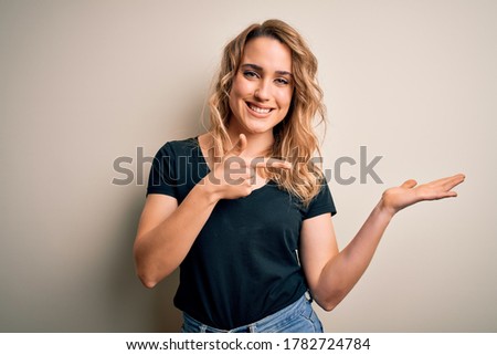 Young beautiful blonde woman wearing casual t-shirt standing over isolated white background amazed and smiling to the camera while presenting with hand and pointing with finger.