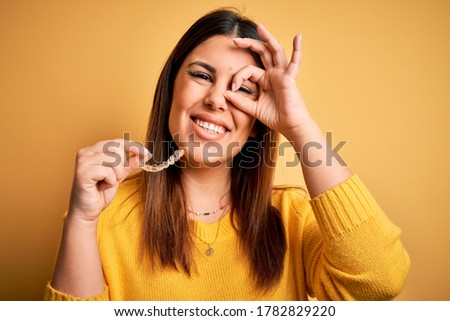 Young beautiful woman holding dental orthodontic aligner corrector over yellow background with happy face smiling doing ok sign with hand on eye looking through fingers