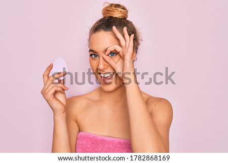 Beautiful woman with blue eyes wearing towel shower after bath holding makeup sponge with happy face smiling doing ok sign with hand on eye looking through fingers