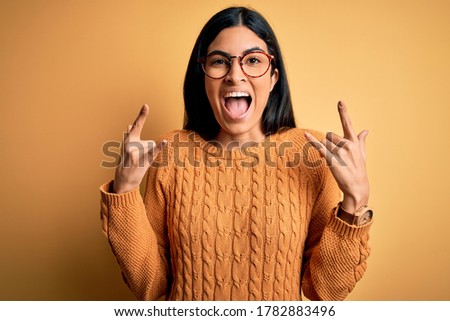 Young beautiful hispanic woman wearing glasses over yellow isolated background shouting with crazy expression doing rock symbol with hands up. Music star. Heavy concept.