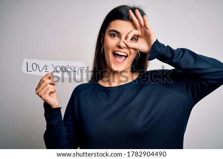 Young beautiful woman asking for take care of you holding paper with love yourself message with happy face smiling doing ok sign with hand on eye looking through fingers