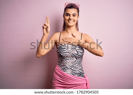 Young beautiful brunette woman on vacation wearing swimsuit over pink background smiling swearing with hand on chest and fingers up, making a loyalty promise oath