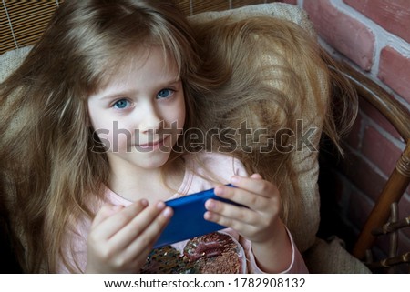 a little blonde girl is watching a video on her phone while sitting in a chair. looks into the frame. Blue eyes